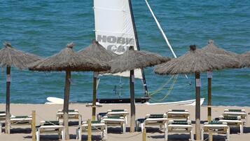 A beach with thatched umbrellas and lounge chairs, a sailboat, and a sandy shore.