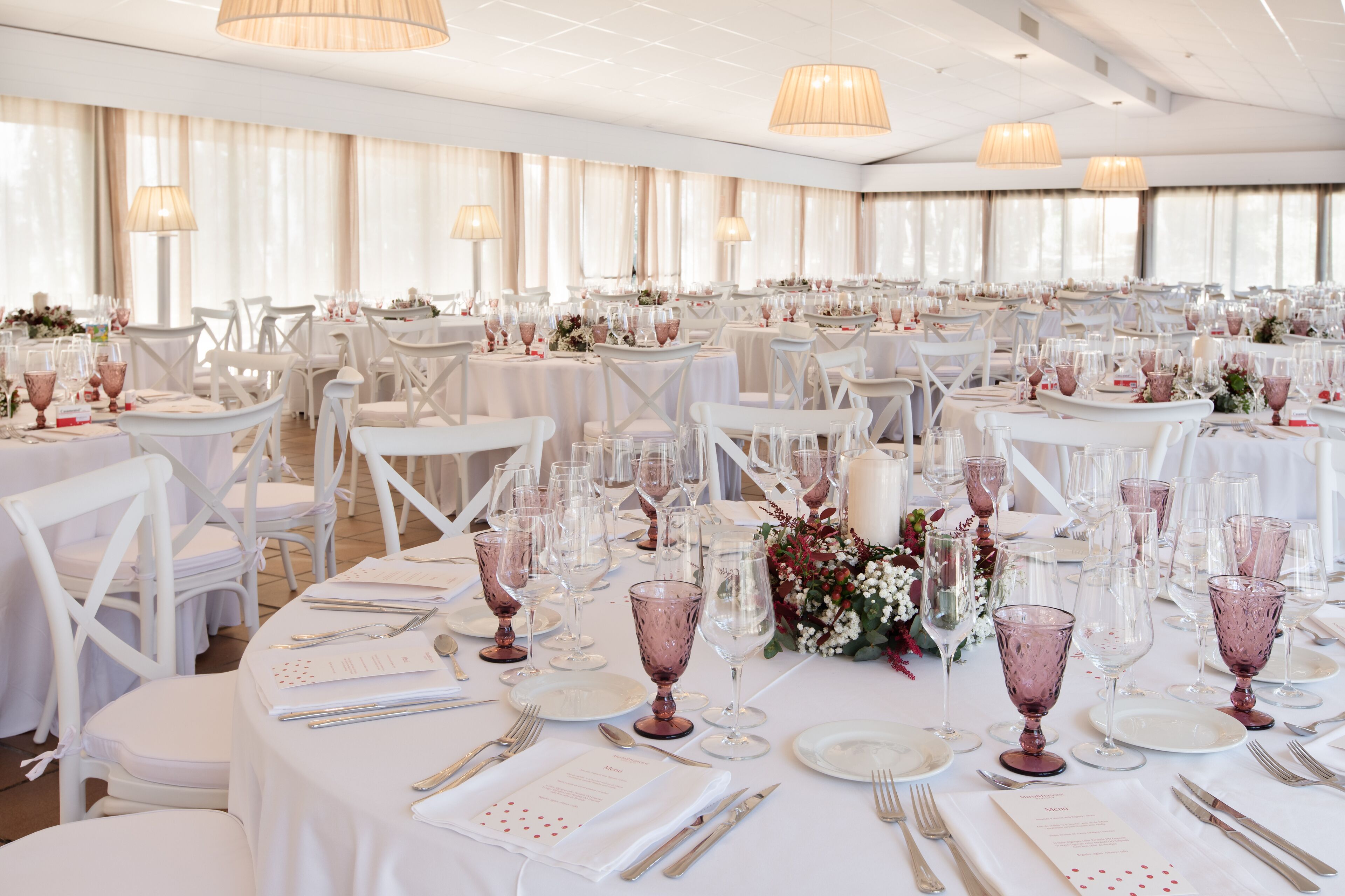 Un salón de banquetes con mesas redondas dispuestas para un evento formal, con sillas blancas, cristalería y centros de mesa florales.