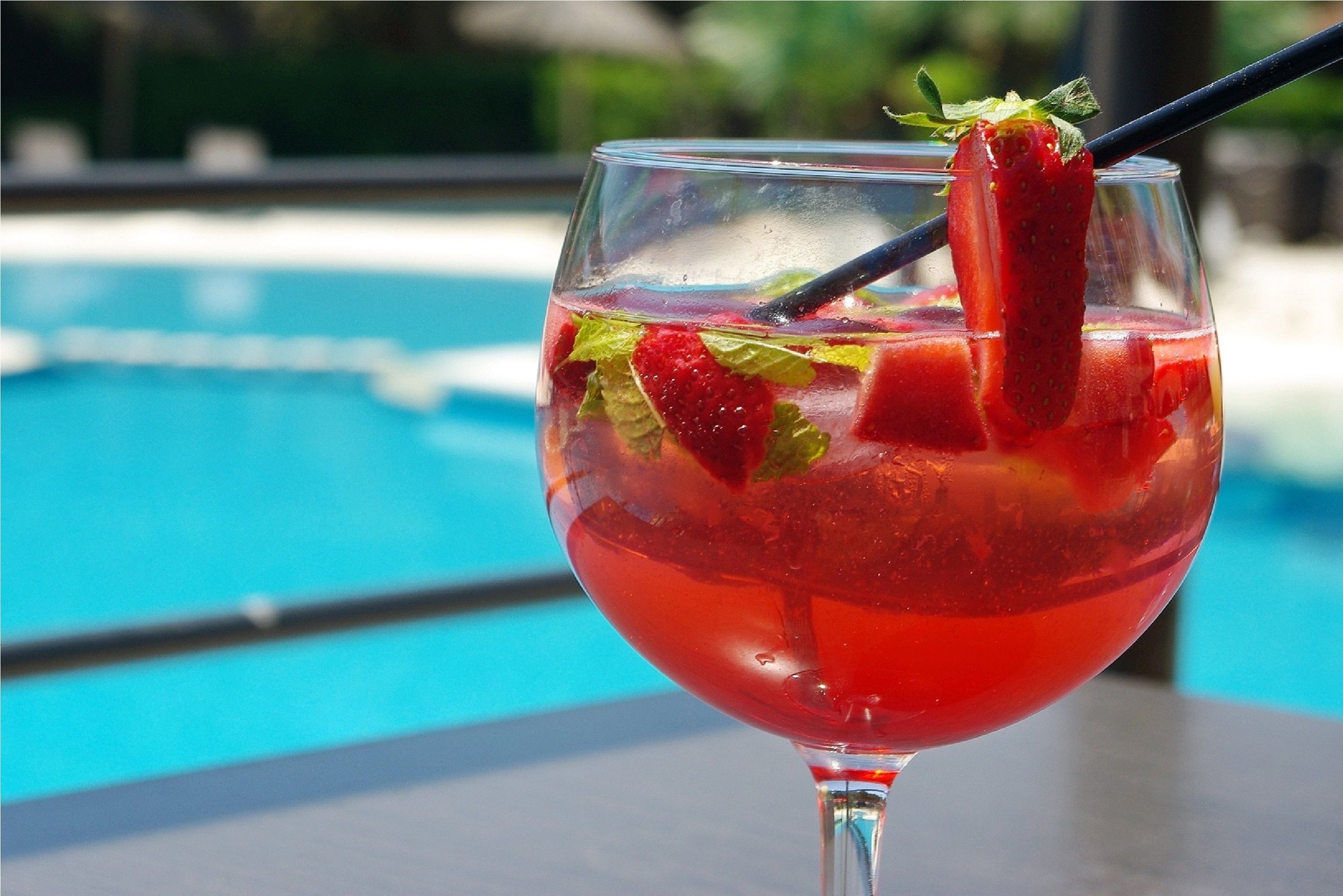 A glass of red beverage with strawberries and mint, placed on a table near a pool.