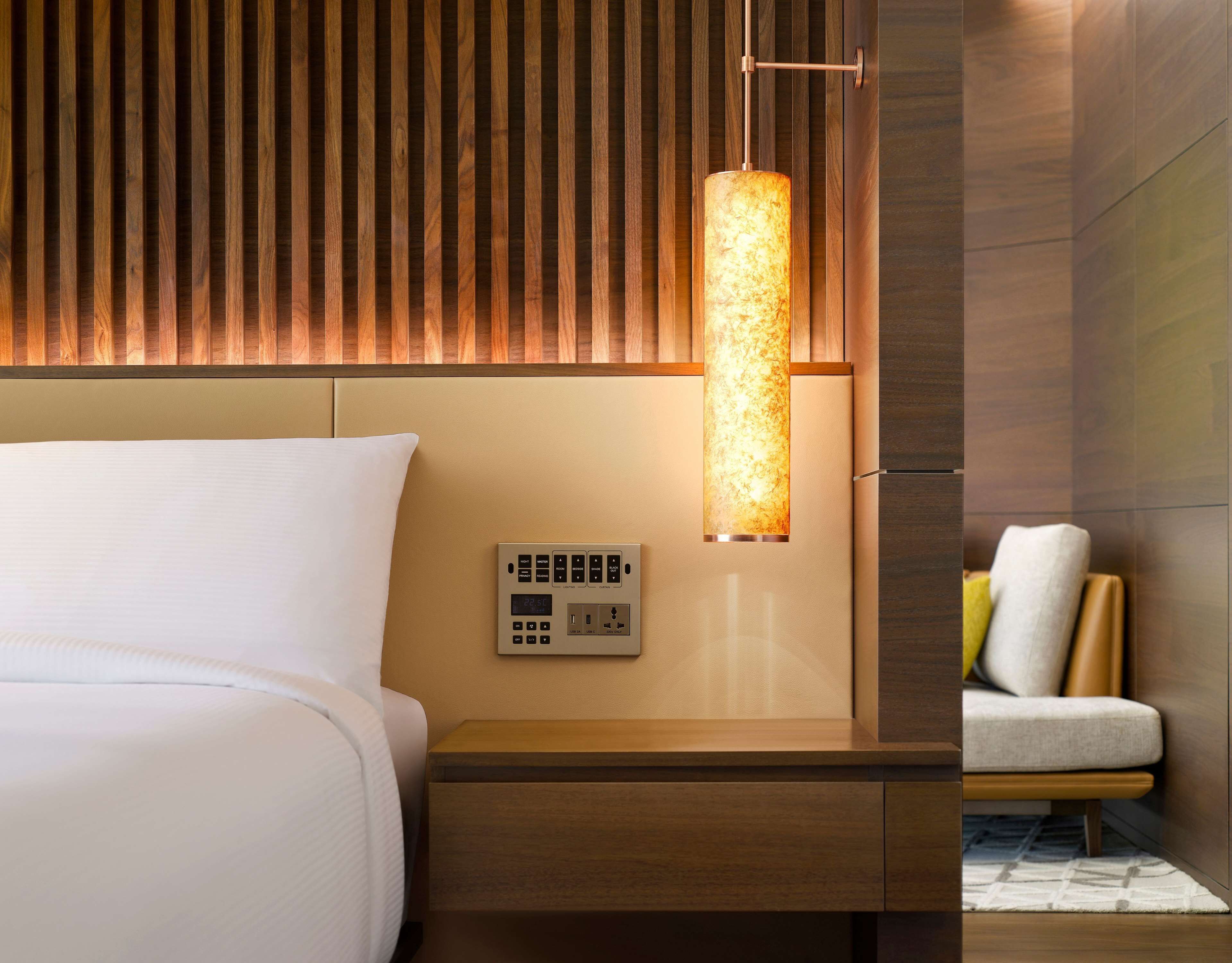 A modern hotel room with a wooden slat headboard, a bedside table with a control panel, and a nightstand.