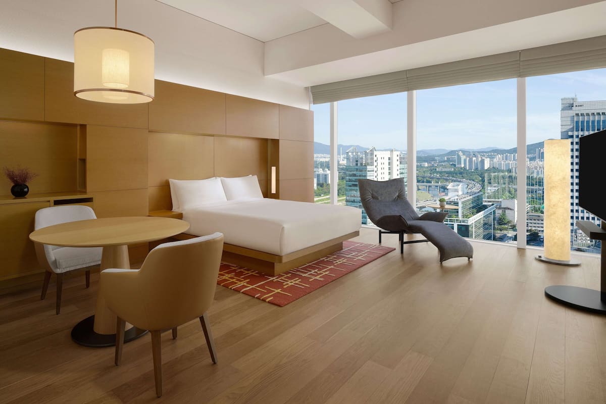 Quarto de hotel moderno com uma cama grande, uma mesa de jantar, duas cadeiras, um sofá e uma TV. O quarto possui amplas janelas com vista para a cidade.