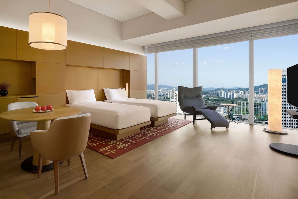 Quarto de hotel moderno com duas camas, uma mesa de jantar, uma cadeira e uma grande janela com vista para a cidade.