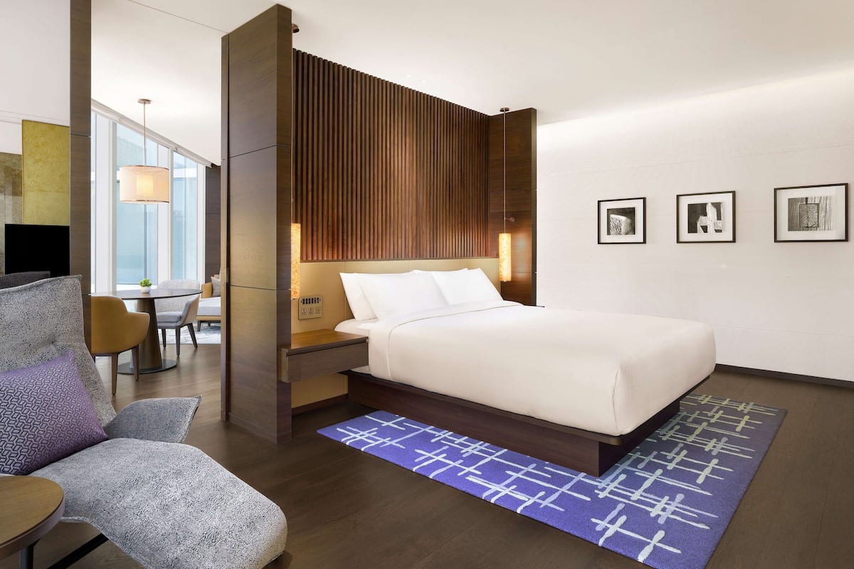 Quarto de hotel moderno com uma cama grande, um espaço de estar com mesa e cadeiras, e vista para a cidade através de amplas janelas.