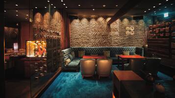 A dimly lit lounge area with a patterned wall, a cushioned seating area, and a bar with glasses on display.
