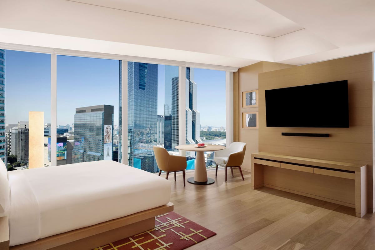 Quarto de hotel moderno com uma cama grande, uma mesa de jantar com cadeiras, uma TV fixada na parede e vista para a cidade através de grandes janelas.