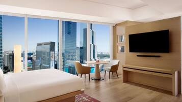 A modern hotel room with a large bed, a dining table with chairs, a TV mounted on the wall, and a city view through large windows.