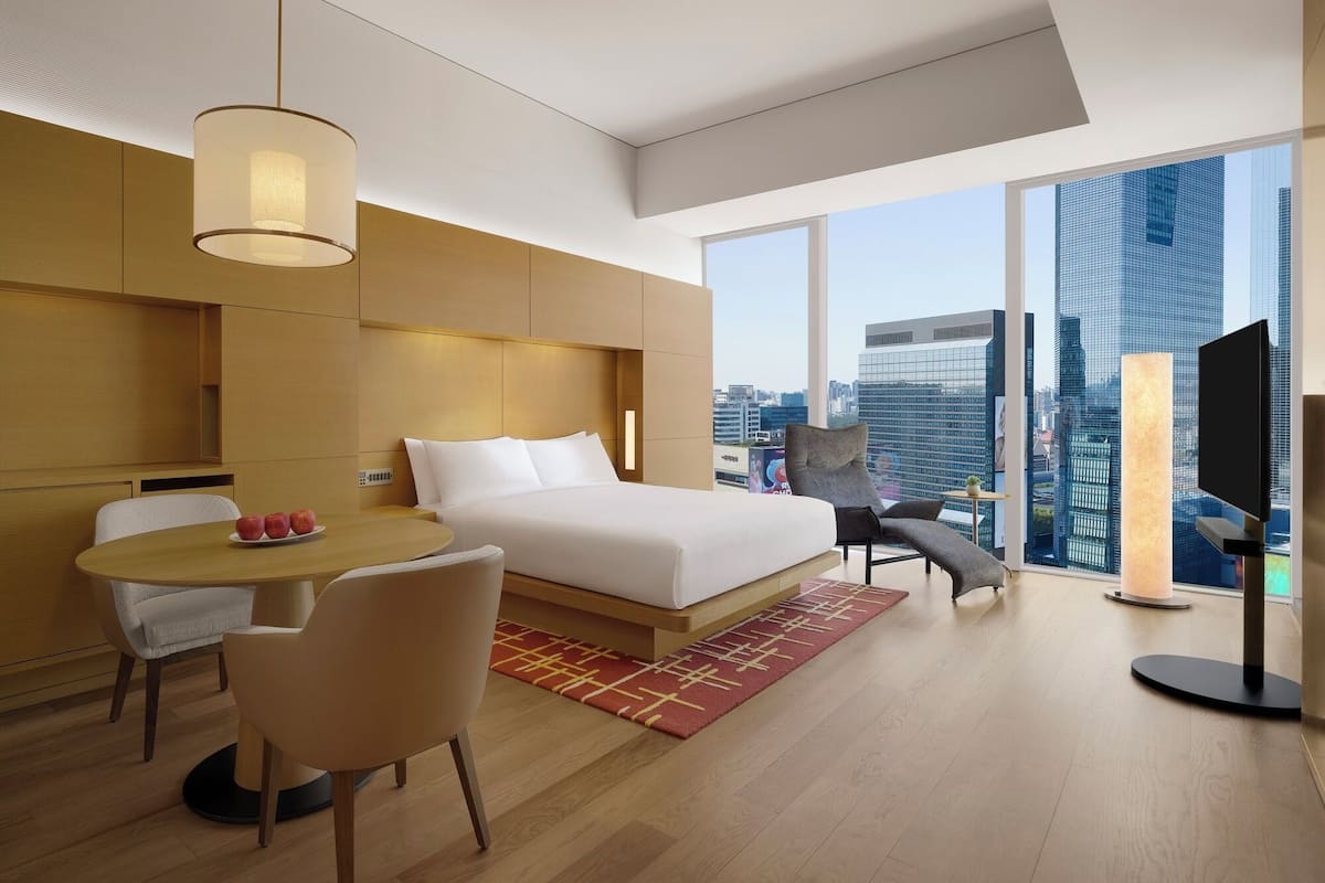 Quarto de hotel moderno com cama, escrivaninha e cadeira. Há uma janela grande com vista para a cidade.