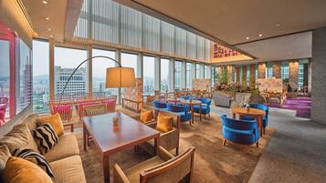 A spacious lounge area with high ceilings, large windows offering city views, and a variety of seating options including sofas and chairs.