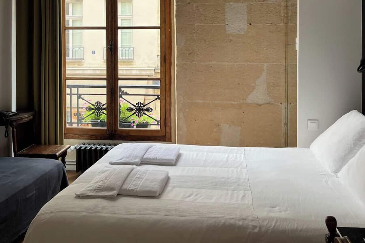 A hotel room with a large bed, a window with a view of a building, a radiator, and a wooden ceiling.