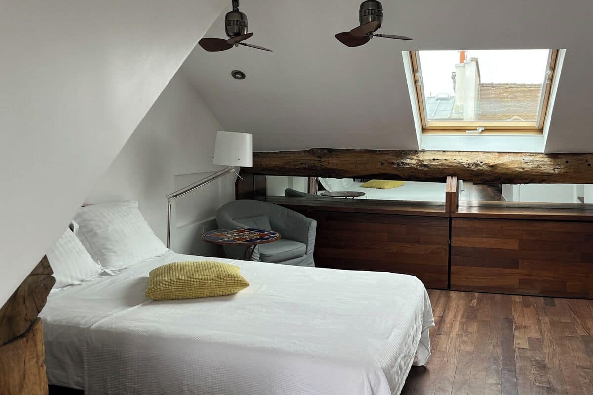 A bedroom with a sloped ceiling, a skylight, a bed with white bedding, a gray armchair, and a wooden desk.