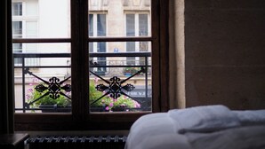 Una habitación con ventana que da a un balcón con baranda de hierro forjado y plantas en macetas.