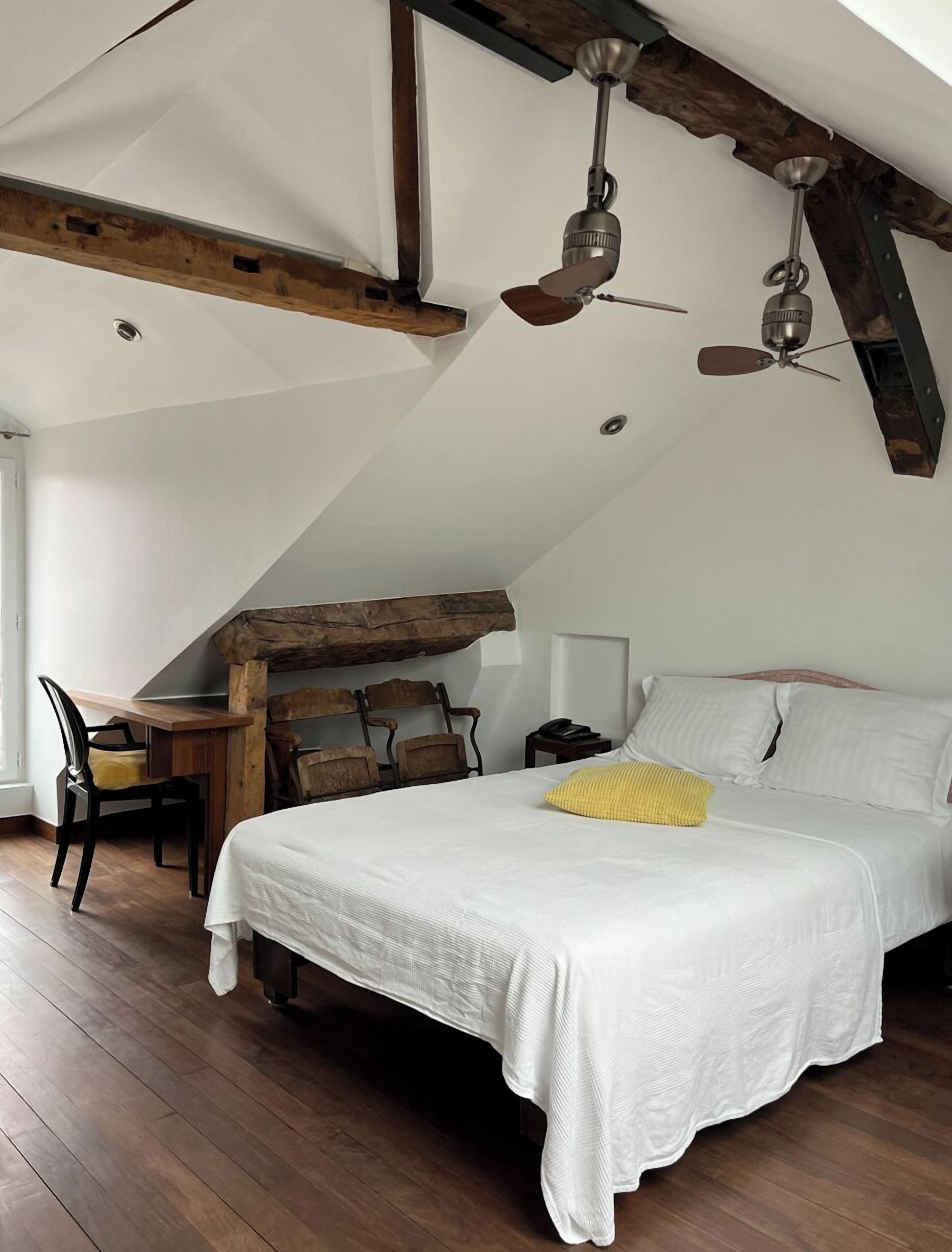 Une chambre avec un plafond en pente, des poutres en bois, un lit avec une literie blanche, un bureau avec une chaise et deux ventilateurs de plafond.