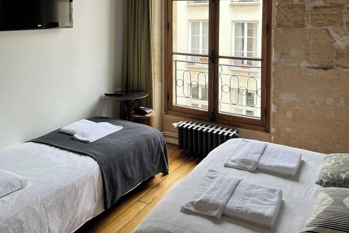 A room with two beds, a television, a window with a view of buildings, and a ceiling fan.
