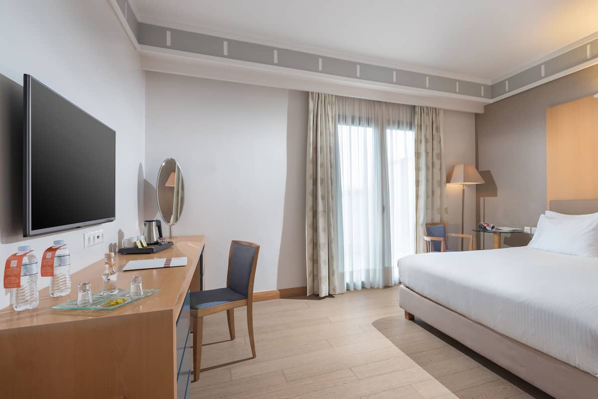 Habitación de hotel con cama, escritorio, televisor de pantalla plana, silla, lámpara y ventana con cortinas.