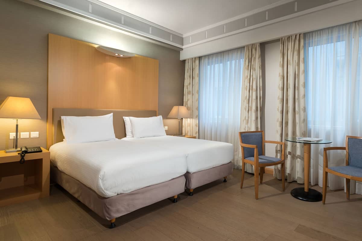 Habitación de hotel con cama doble, dos mesitas de noche con lámparas, una mesita redonda pequeña con dos sillas y un ventanal con cortinas.