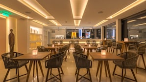 A modern restaurant with wooden tables and chairs, a bar area, and a view of the city through large windows. - Central Athens Hotel (Athens)