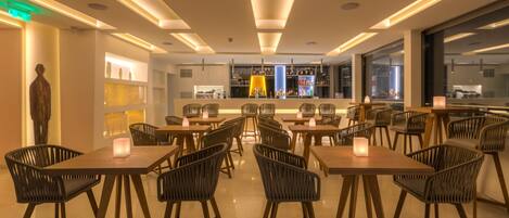 A modern restaurant with wooden tables and chairs, a bar area, and a view of the city through large windows.