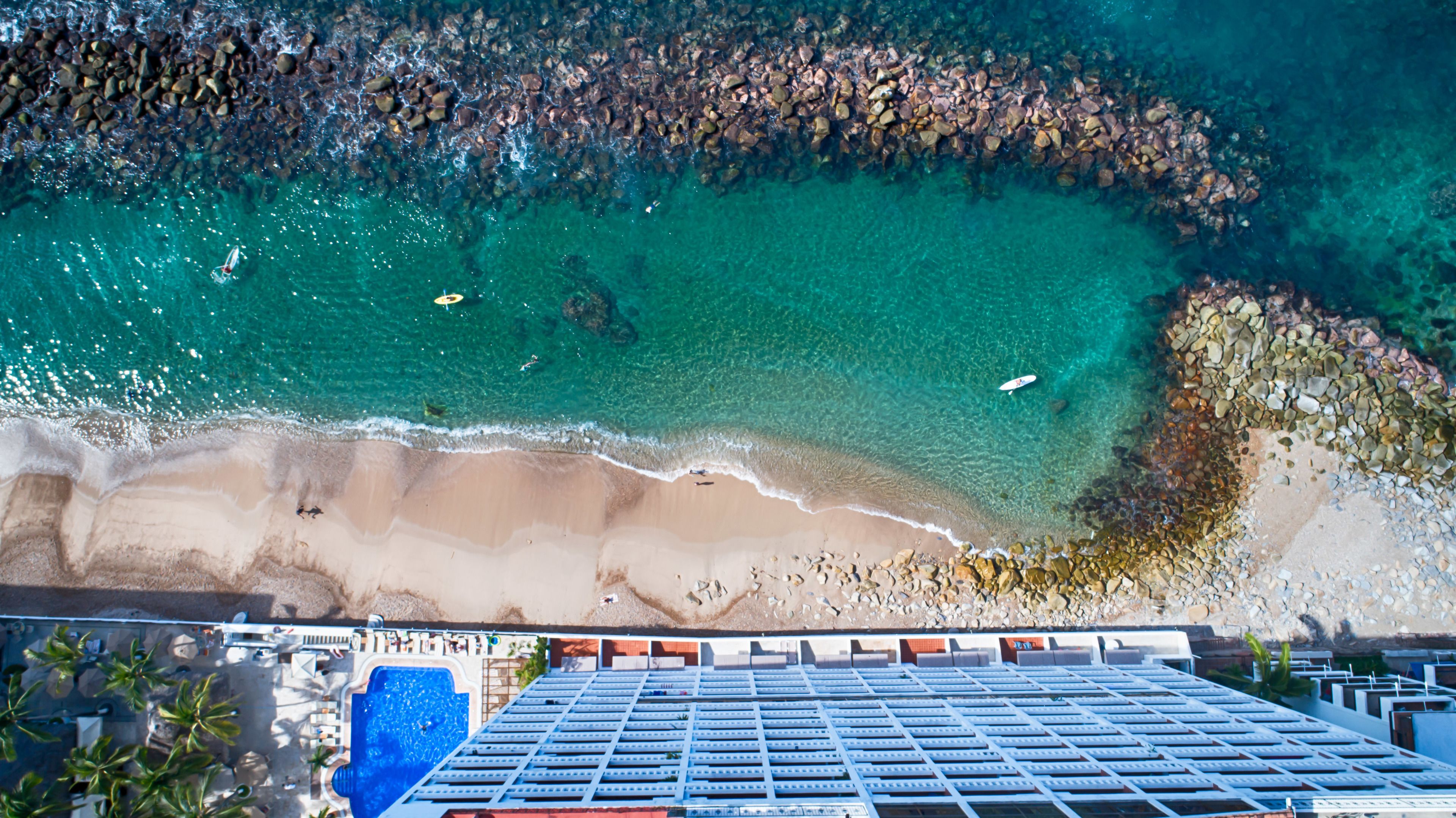 An aerial view of a beach with clear turquoise water, a sandy shore, and a building with a pool in the foreground.