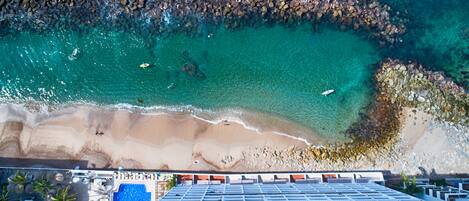 An aerial view of a beach with clear turquoise water, a sandy shore, and a building with a pool in the foreground.