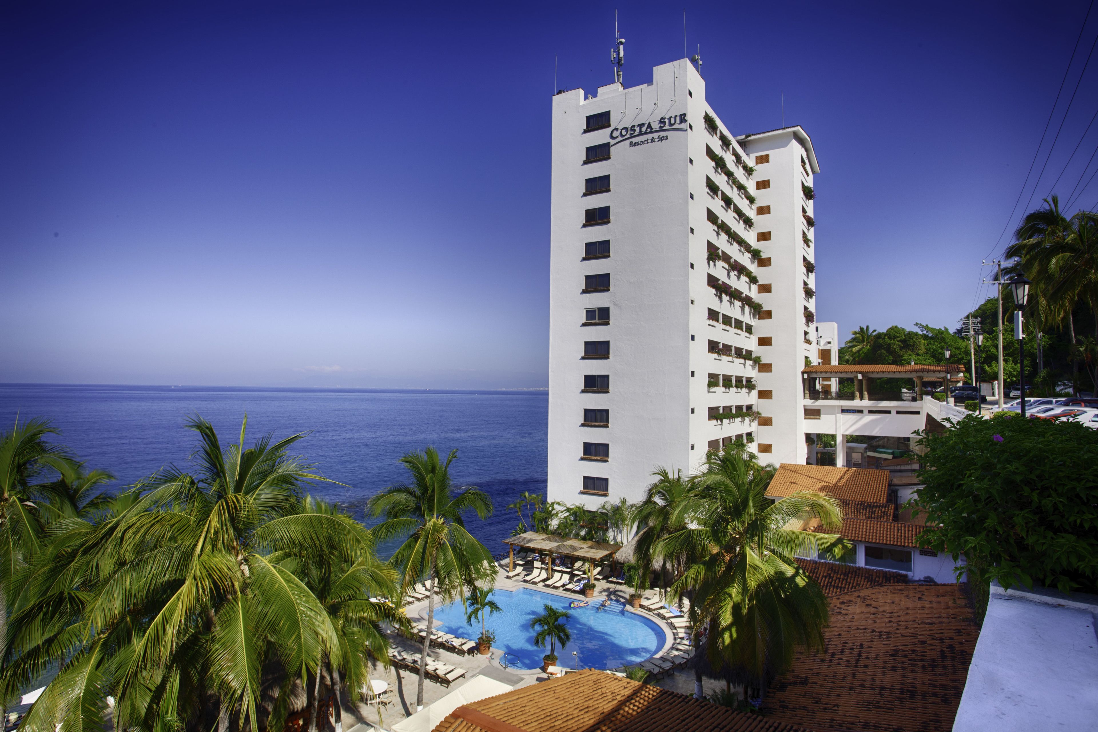 Um hotel moderno com piscina, cercado por palmeiras e vista para o oceano.