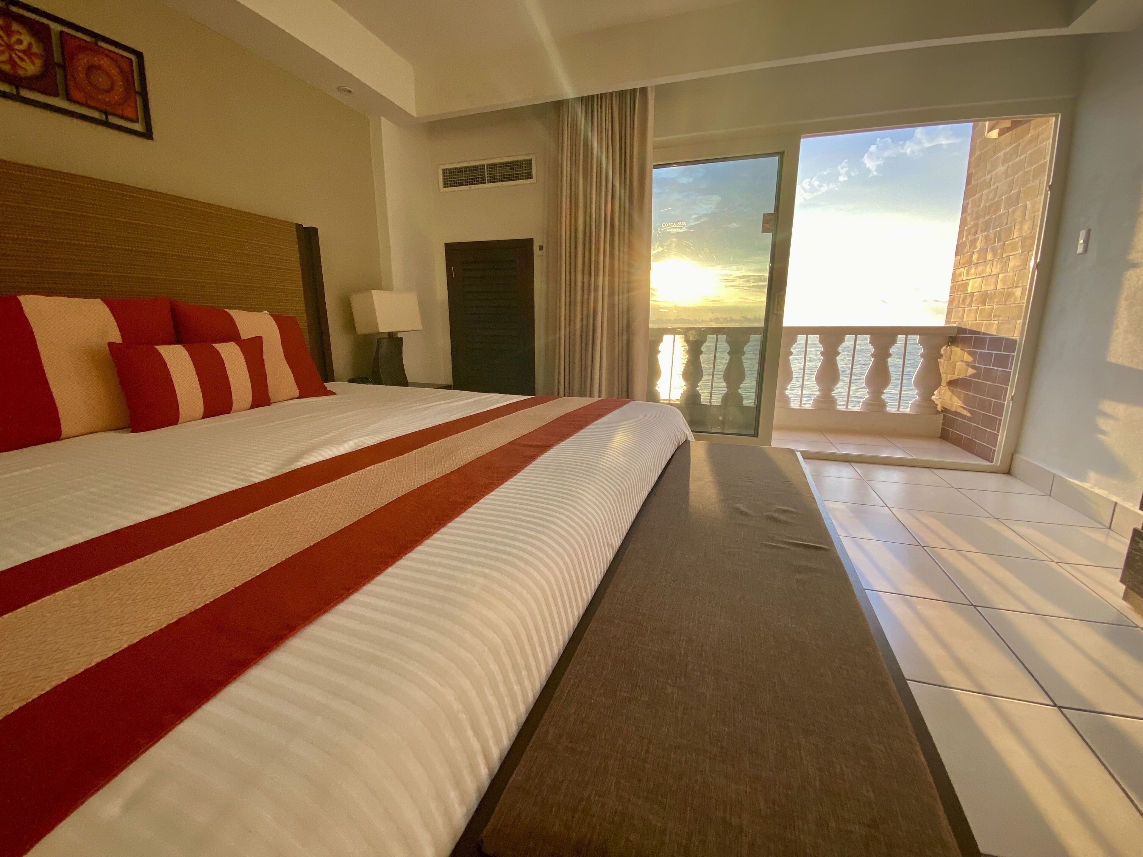 A hotel room with a large bed, a nightstand, a lamp, a view of the sunset through a balcony door, and a tiled floor.