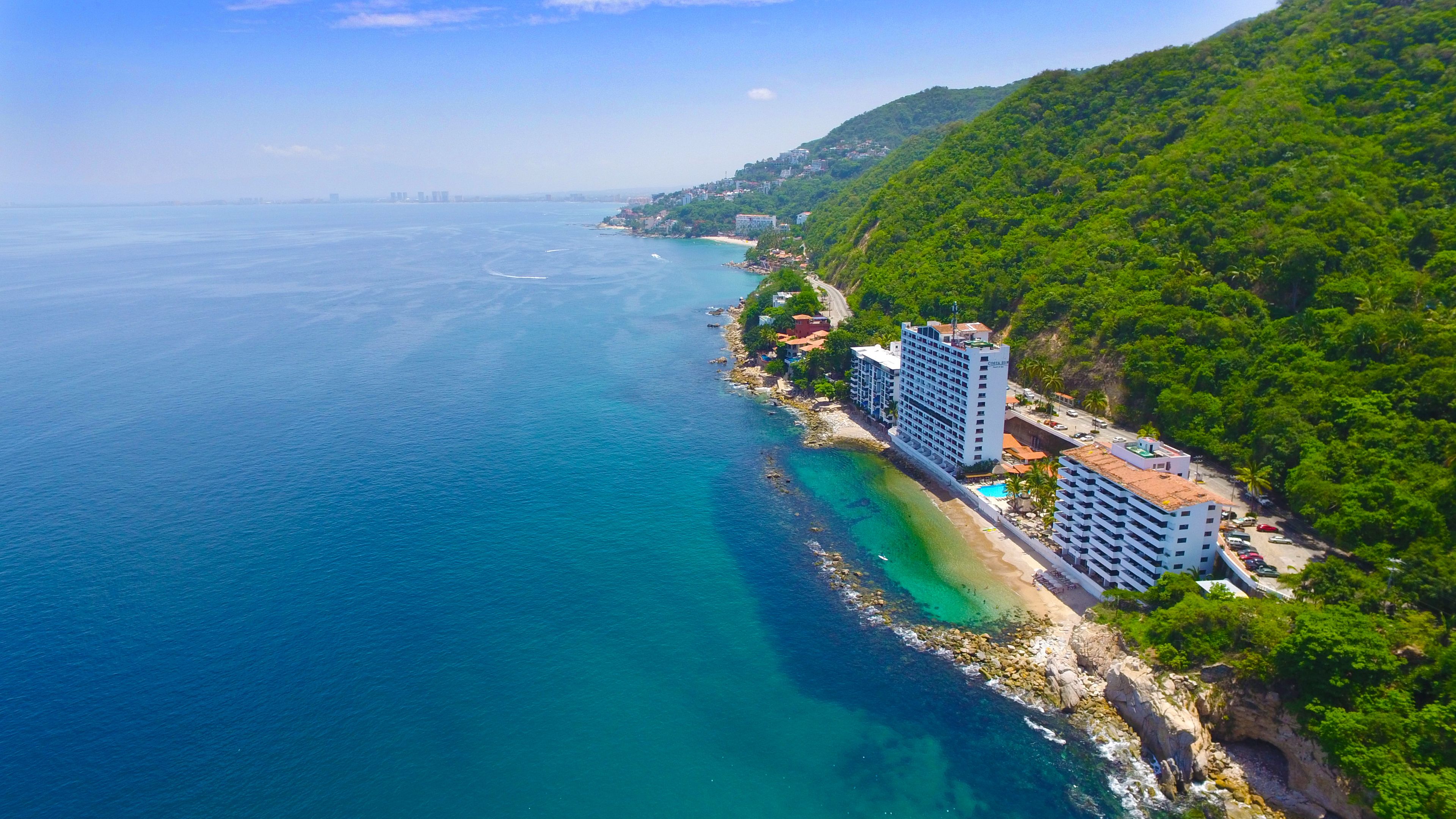 An aerial view of a coastal area with a hotel complex adjacent to a beach, featuring a rocky shoreline and lush greenery.