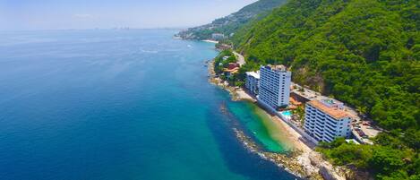 An aerial view of a coastal area with a hotel complex adjacent to a beach, featuring a rocky shoreline and lush greenery.