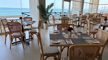 Un restaurante con sillas y mesas de mimbre, ventanas amplias con vistas al mar y plantas en macetas.