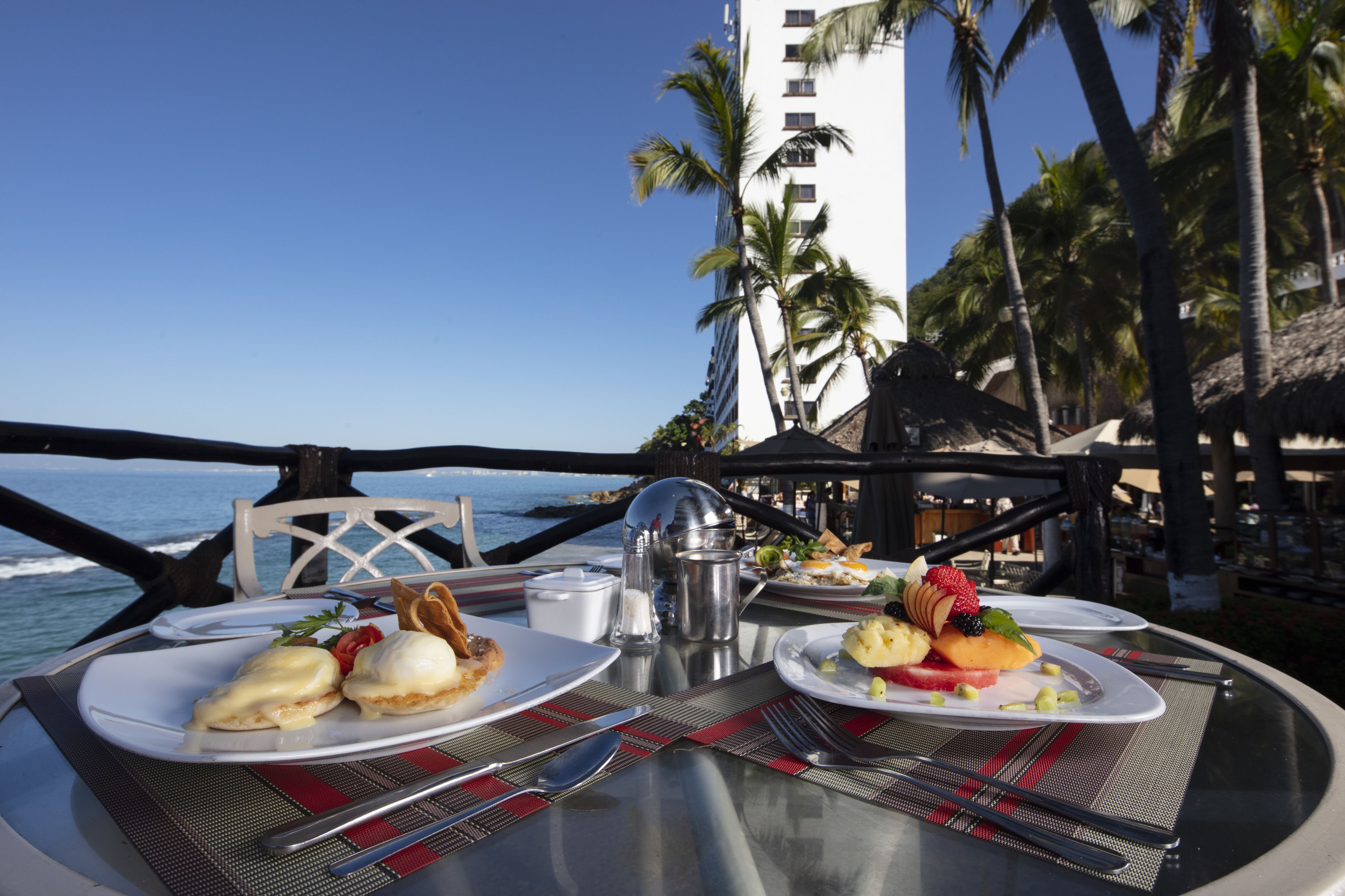 A table set with breakfast dishes overlooking the ocean, palm trees, and a hotel building.