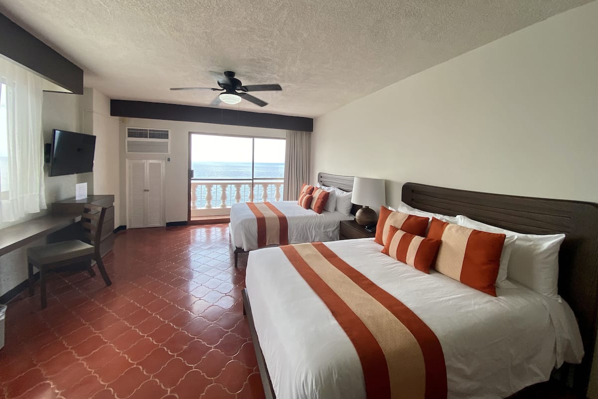 A hotel room with two beds, a desk, a television, and a balcony with a view of the sea.