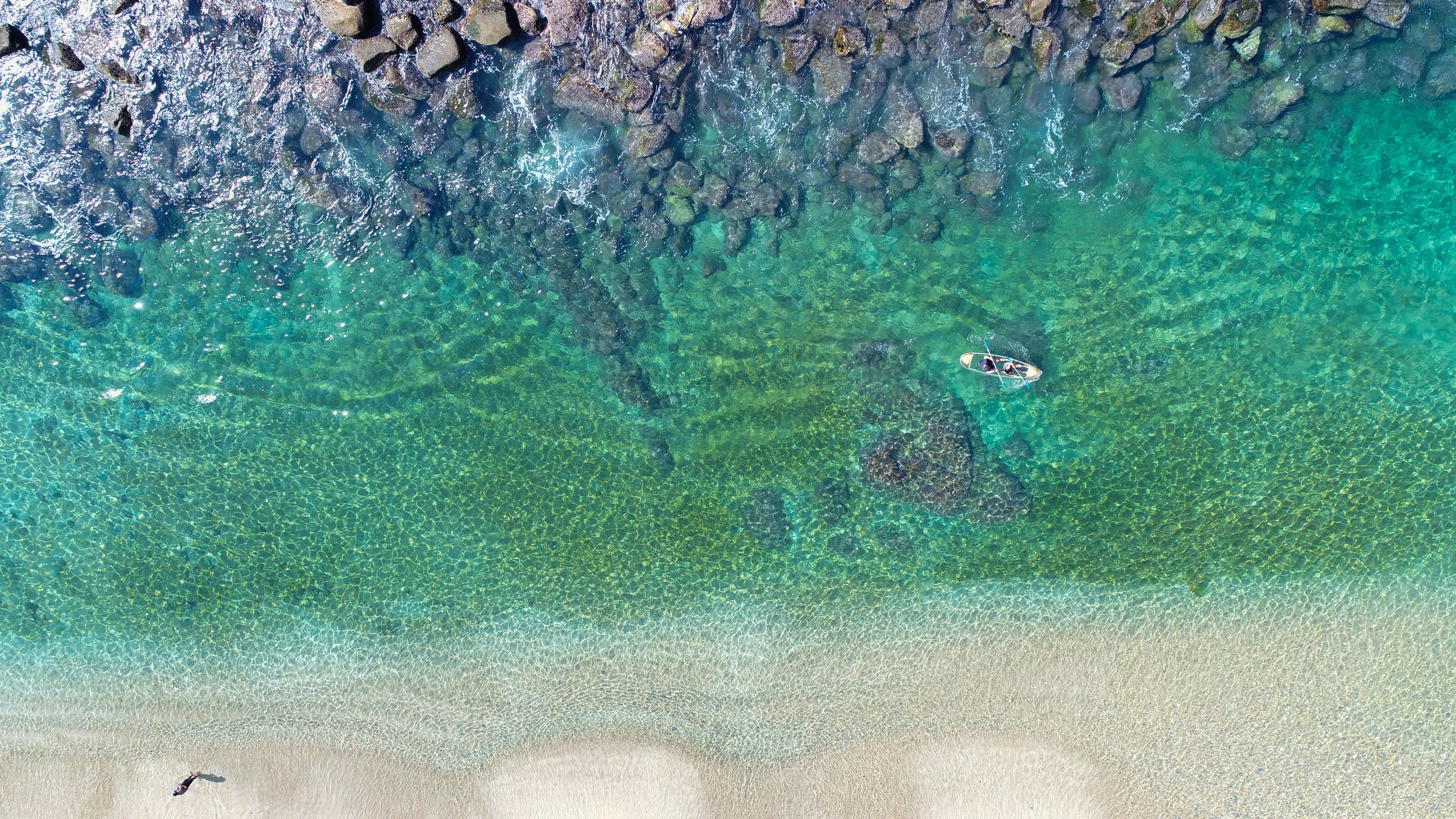 An aerial view of a beach with clear turquoise water, a rocky shoreline, and a person paddling in the water.