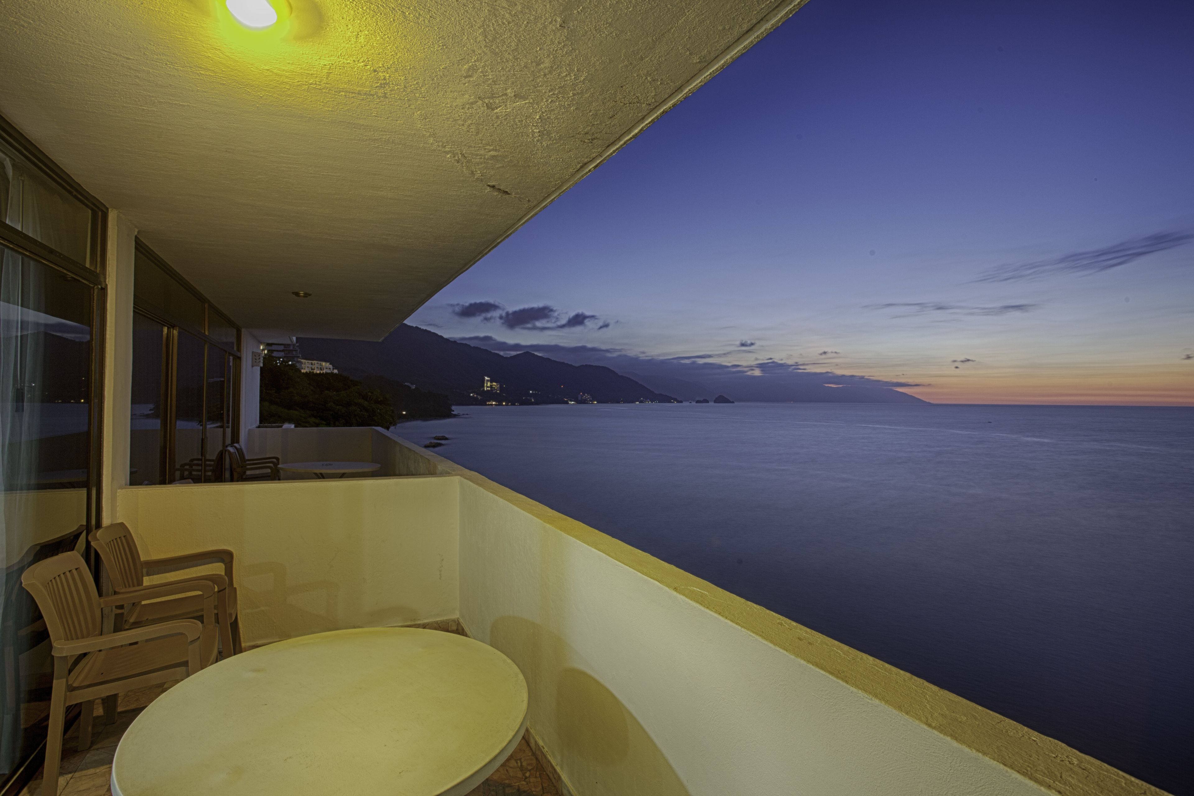 Ein Balkon mit Tisch und Stühlen, der Blick auf ein ruhiges Meer und ferne Berge im Abendlicht gewährt.