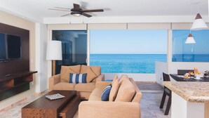 A modern living room with a large TV, comfortable seating, a ceiling fan, and a view of the ocean.