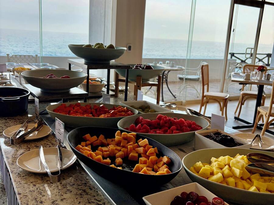Un bufé con diversas frutas y verduras, un comedor con sillas y mesas, y vista al océano a través de amplias ventanas.