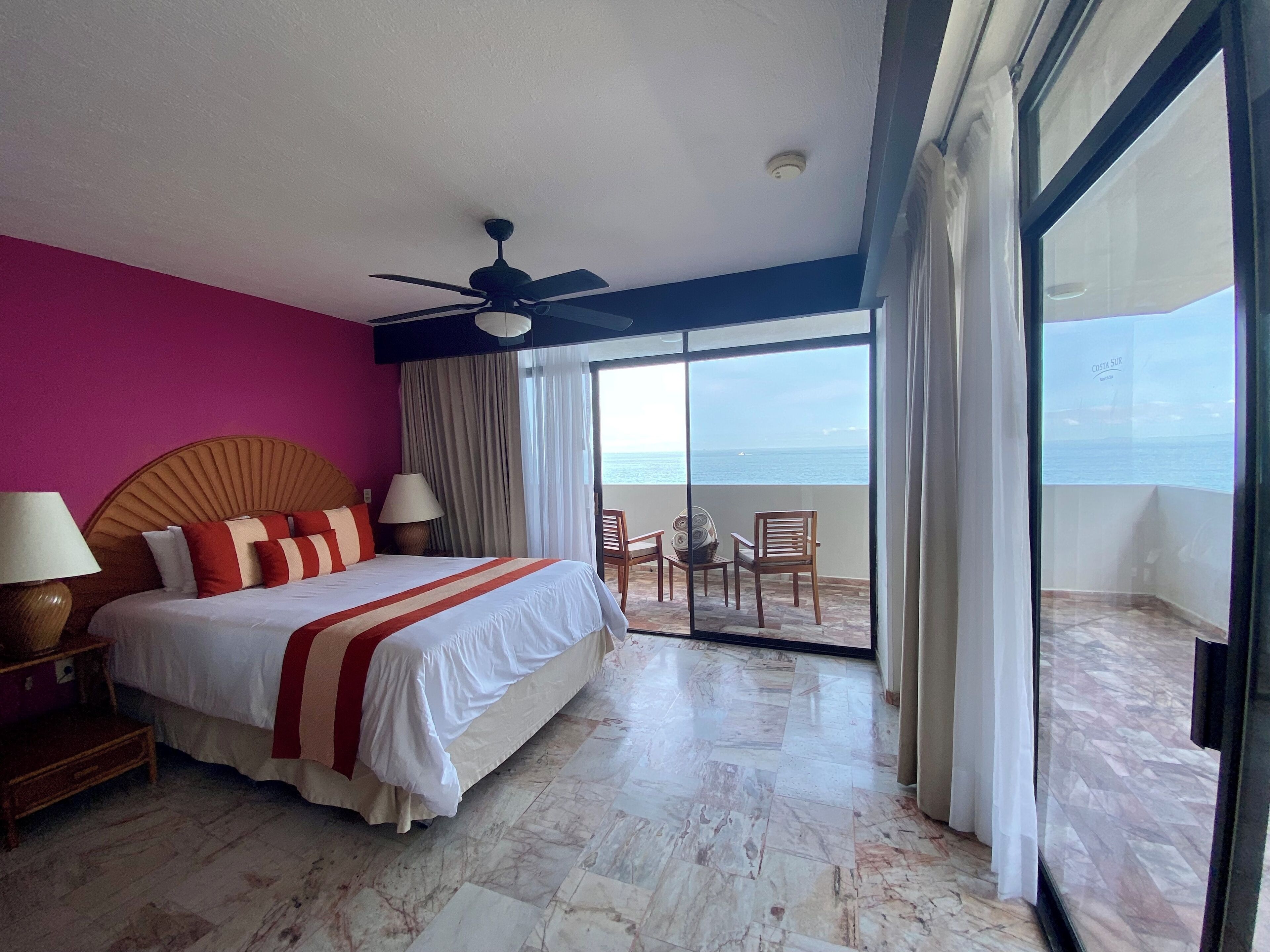 A hotel room with a bed, a ceiling fan, a view of the ocean through large windows, and a balcony with chairs.