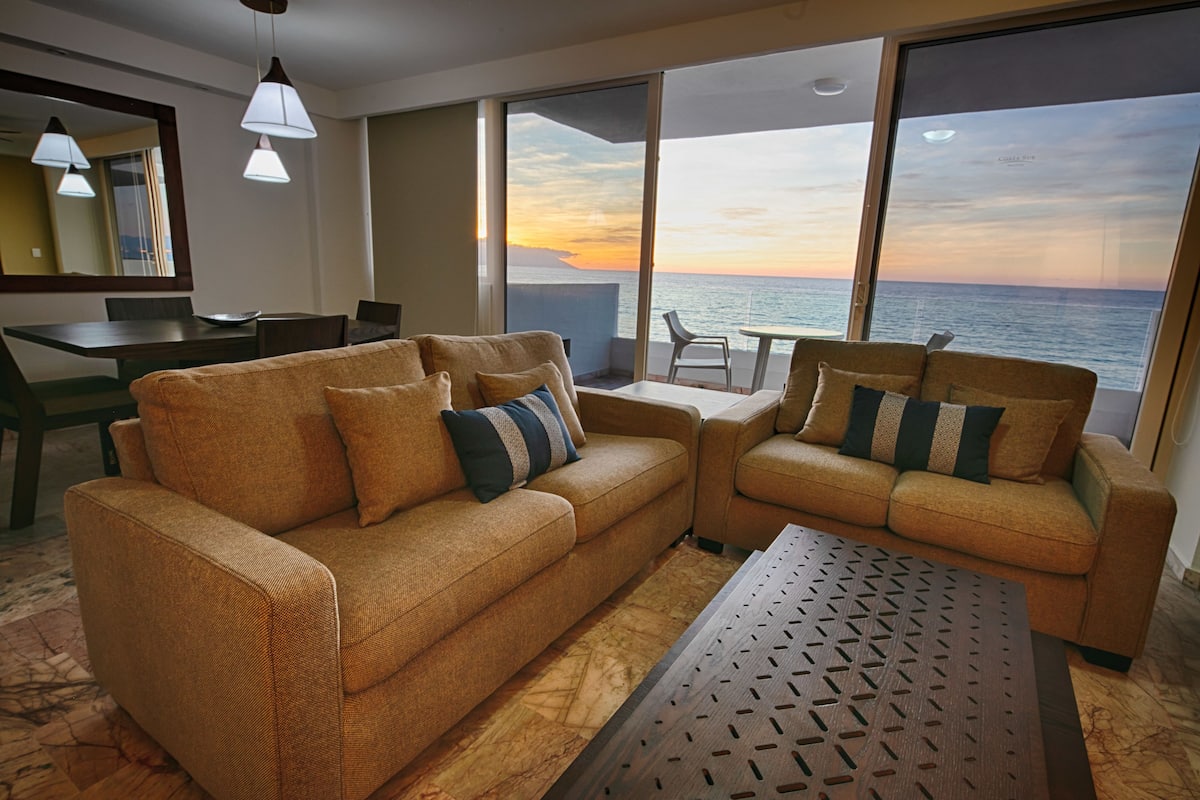 A modern living room with two sofas, a dining table, and a large window with a view of the ocean.