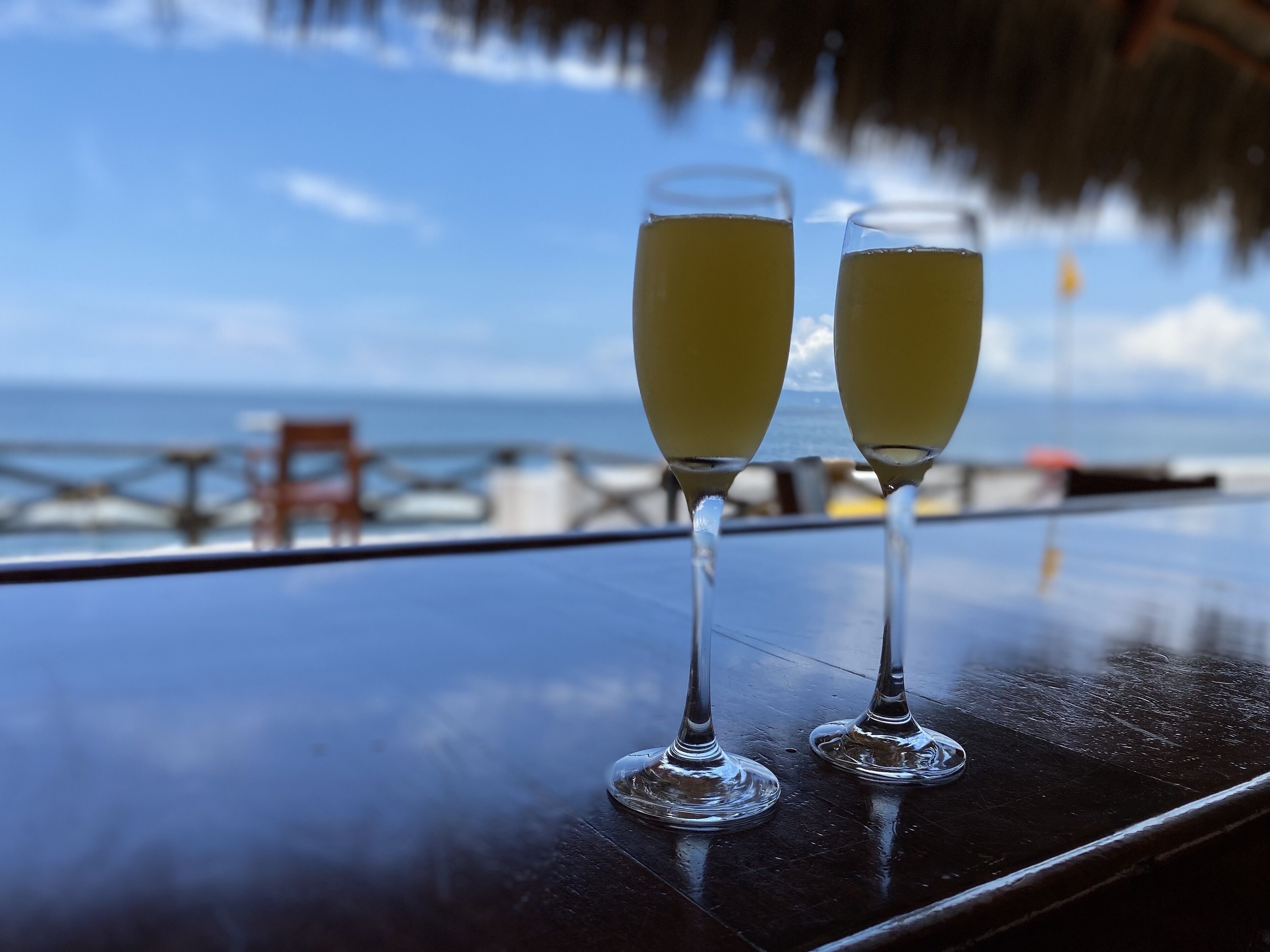 Dos vasos de una bebida amarilla sobre una mesa con vista al océano y una estructura de techo de paja.