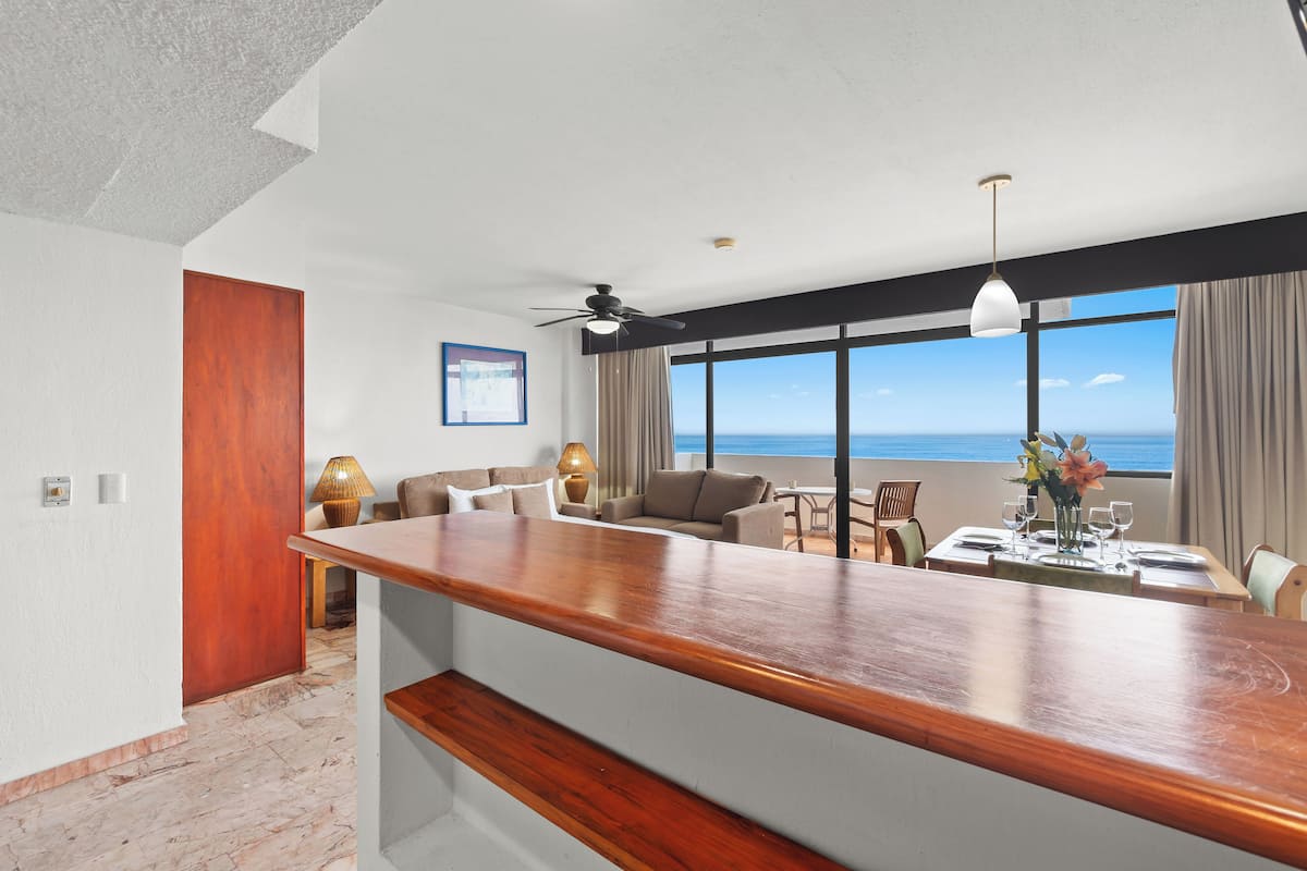A modern hotel room with a wooden bar, a sofa, a dining table set for two, and a large window offering a view of the ocean.
