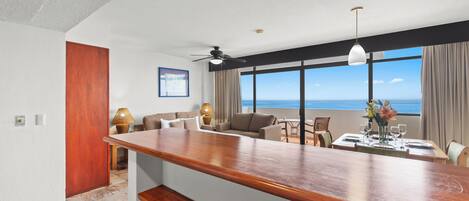 Habitación de hotel moderna con barra de madera, sofá, mesa de comedor para dos personas y un gran ventanal con vista al océano.