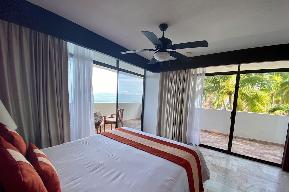 A hotel room with a large bed, a ceiling fan, and a balcony with a view of palm trees and the ocean.