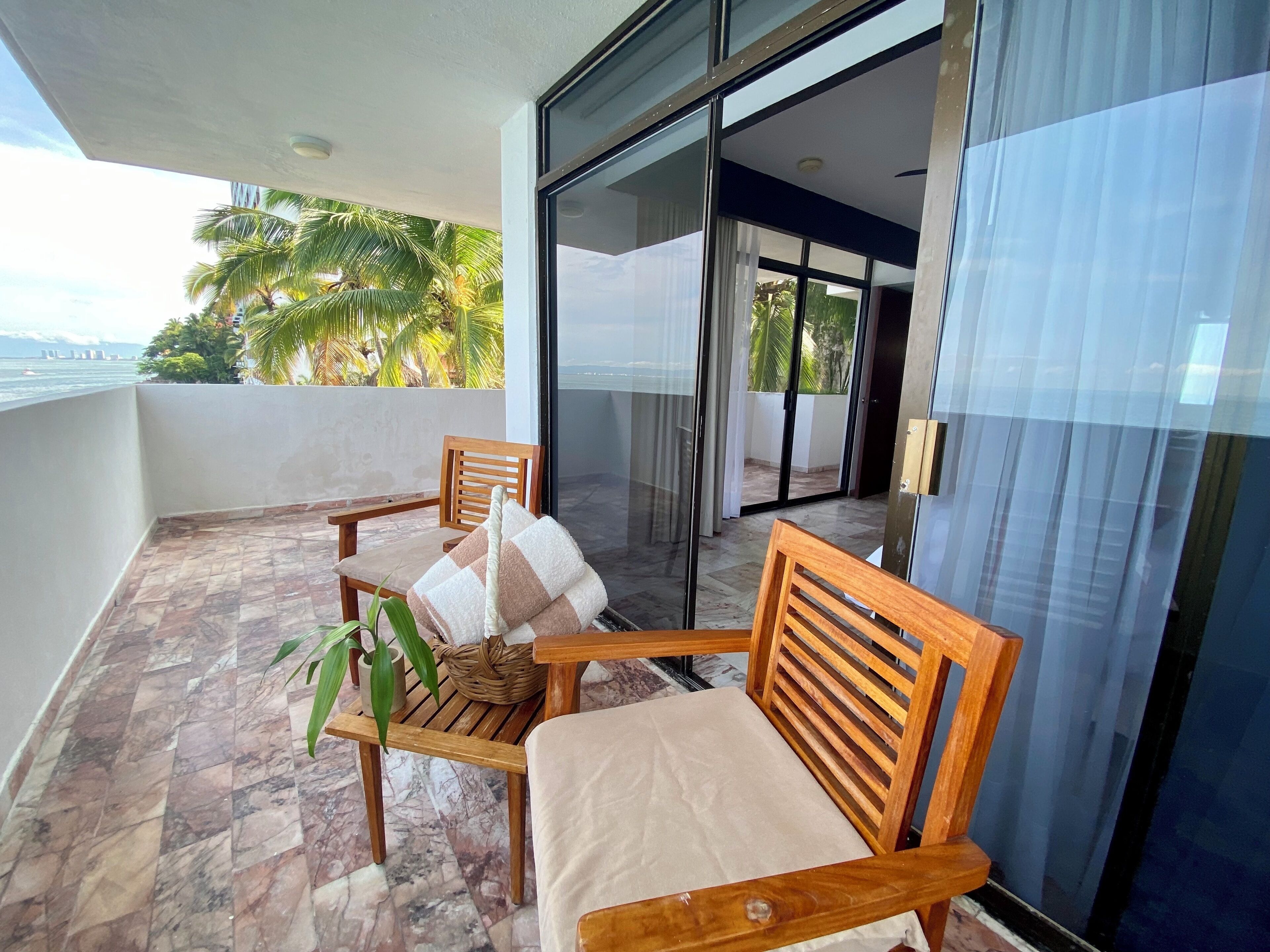 Un balcon avec une chaise en bois et une petite table, un pot de fleurs et une vue sur les palmiers et la mer.