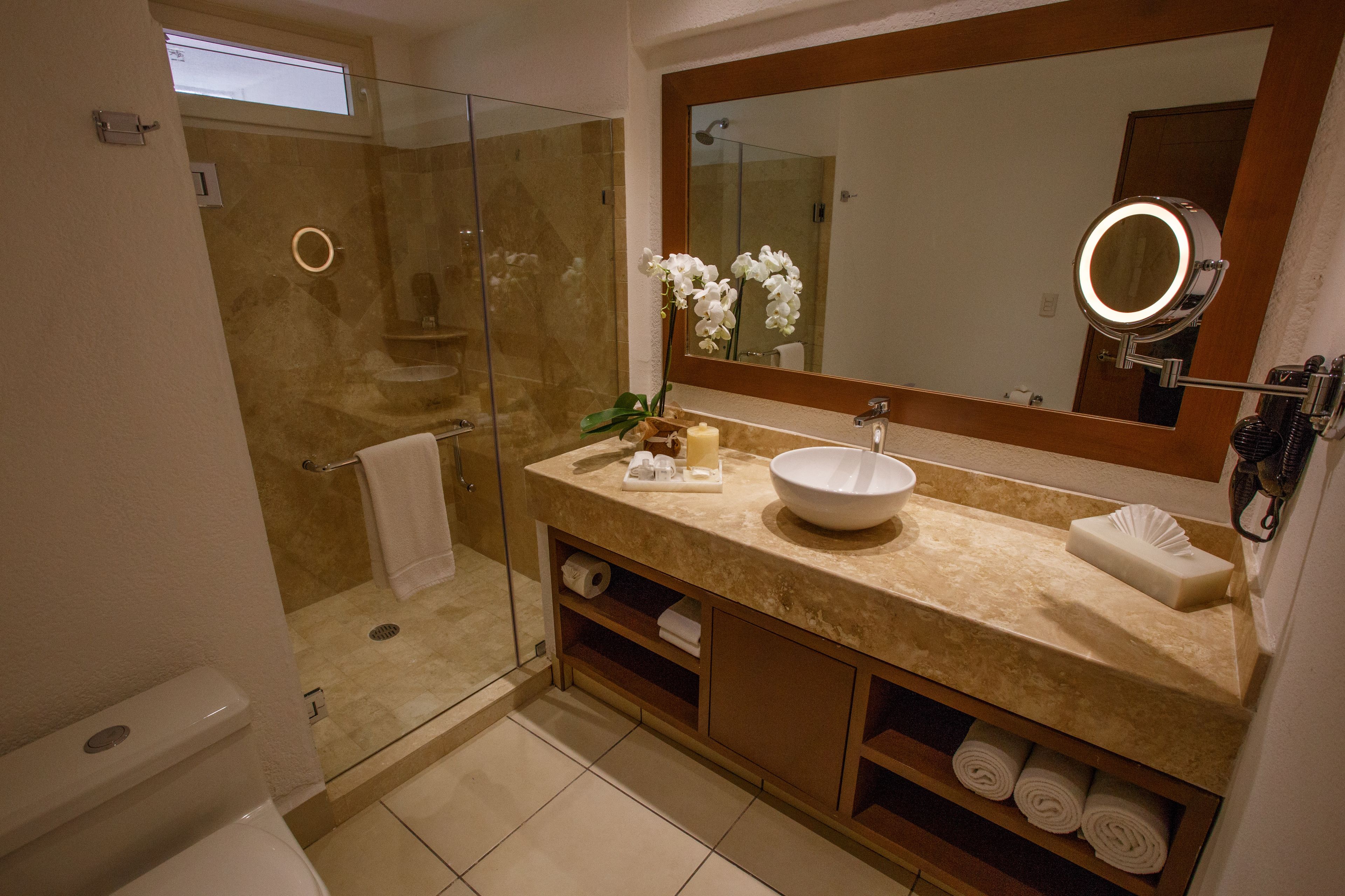 Een moderne badkamer met een douchecabine van glas, een grote spiegel met ronde verlichting en een beige aanrecht met een wastafel en handdoekenkast.