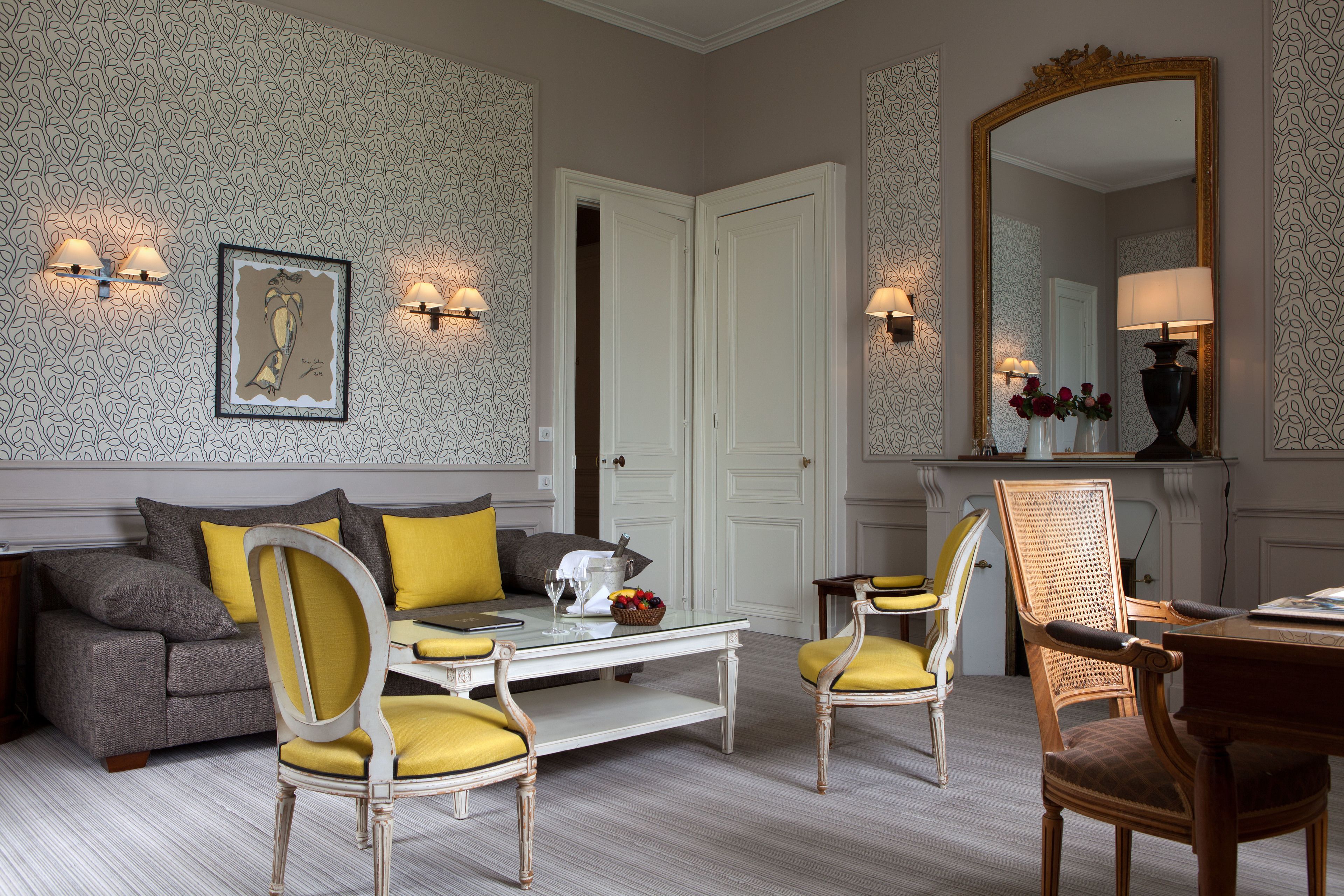 Ein Wohnzimmer mit einer Couch, zwei Sesseln, einem Couchtisch und einem Esstisch. An den Wänden befinden sich Wandleuchten, ein gerahmtes Kunstwerk und ein Spiegel.