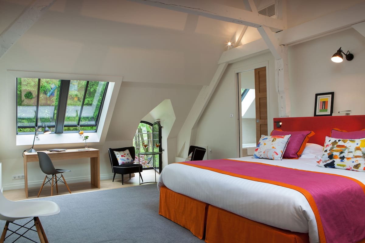 A bedroom with a large bed, a desk, and a chair. There is a window providing a view of the outdoors.