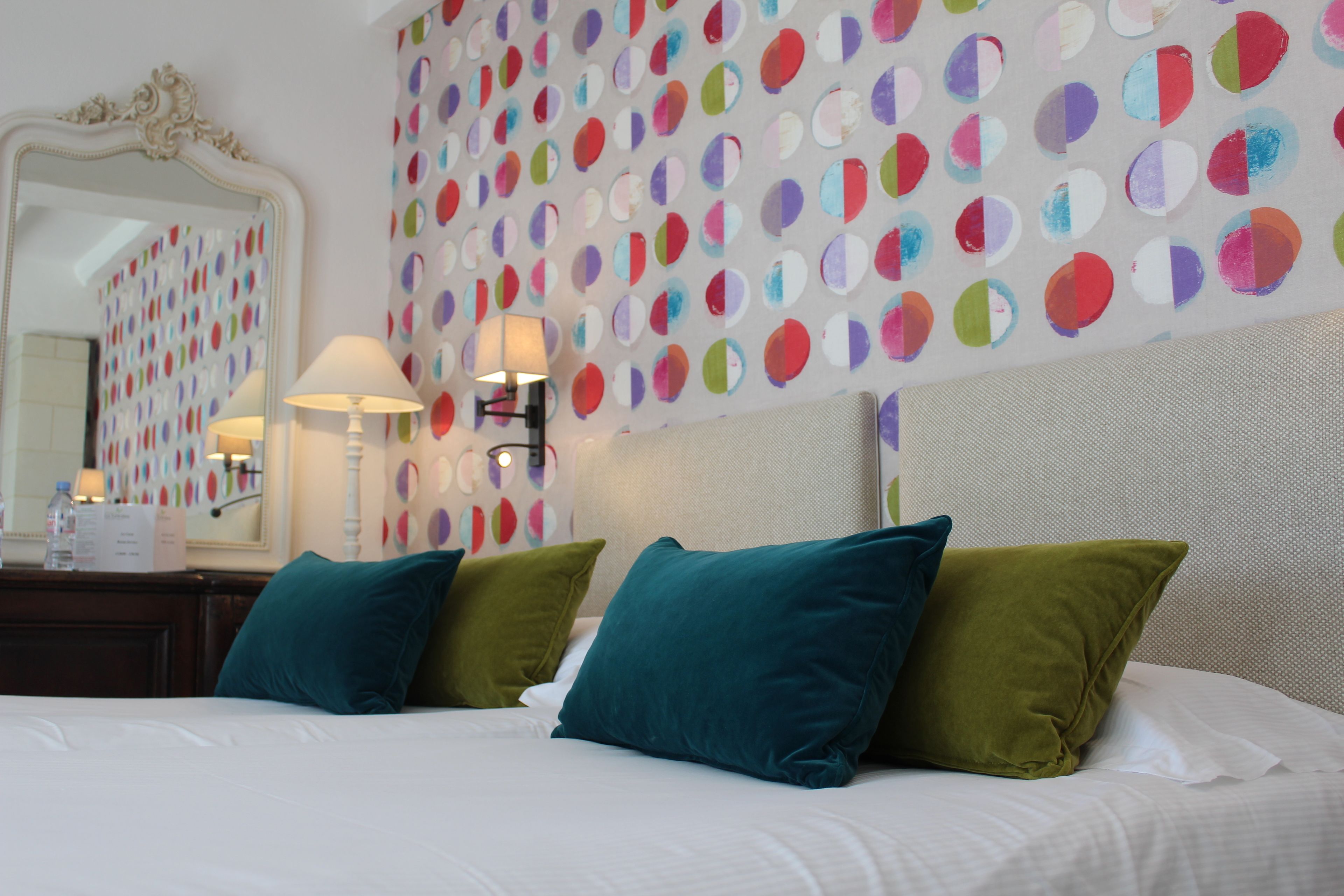 A hotel room with a bed, pillows, a nightstand, a mirror, and a wall with colorful circular patterns.