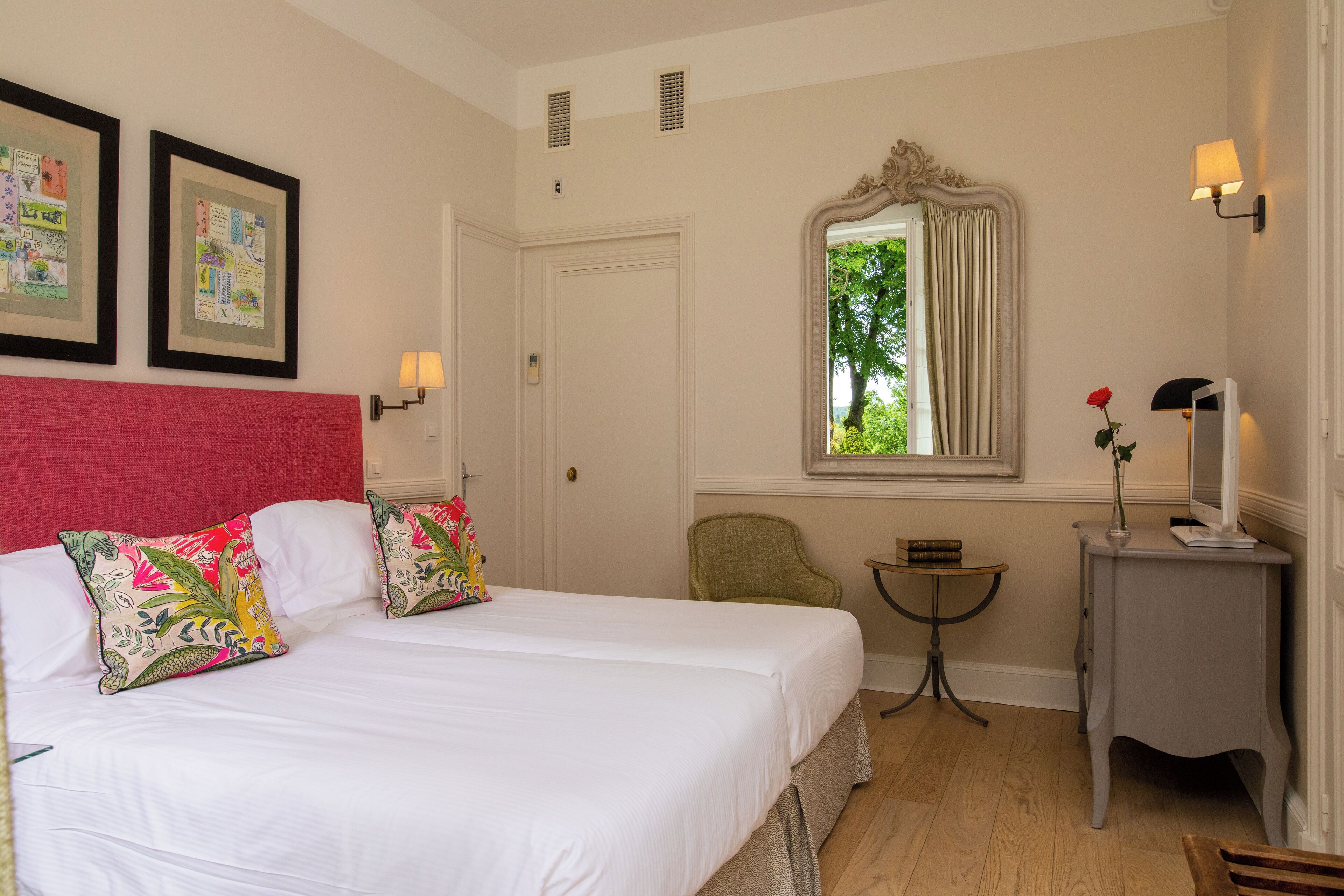 A hotel room with a large bed, two framed pictures on the wall, a window with a view of greenery, a small table, and a chair.