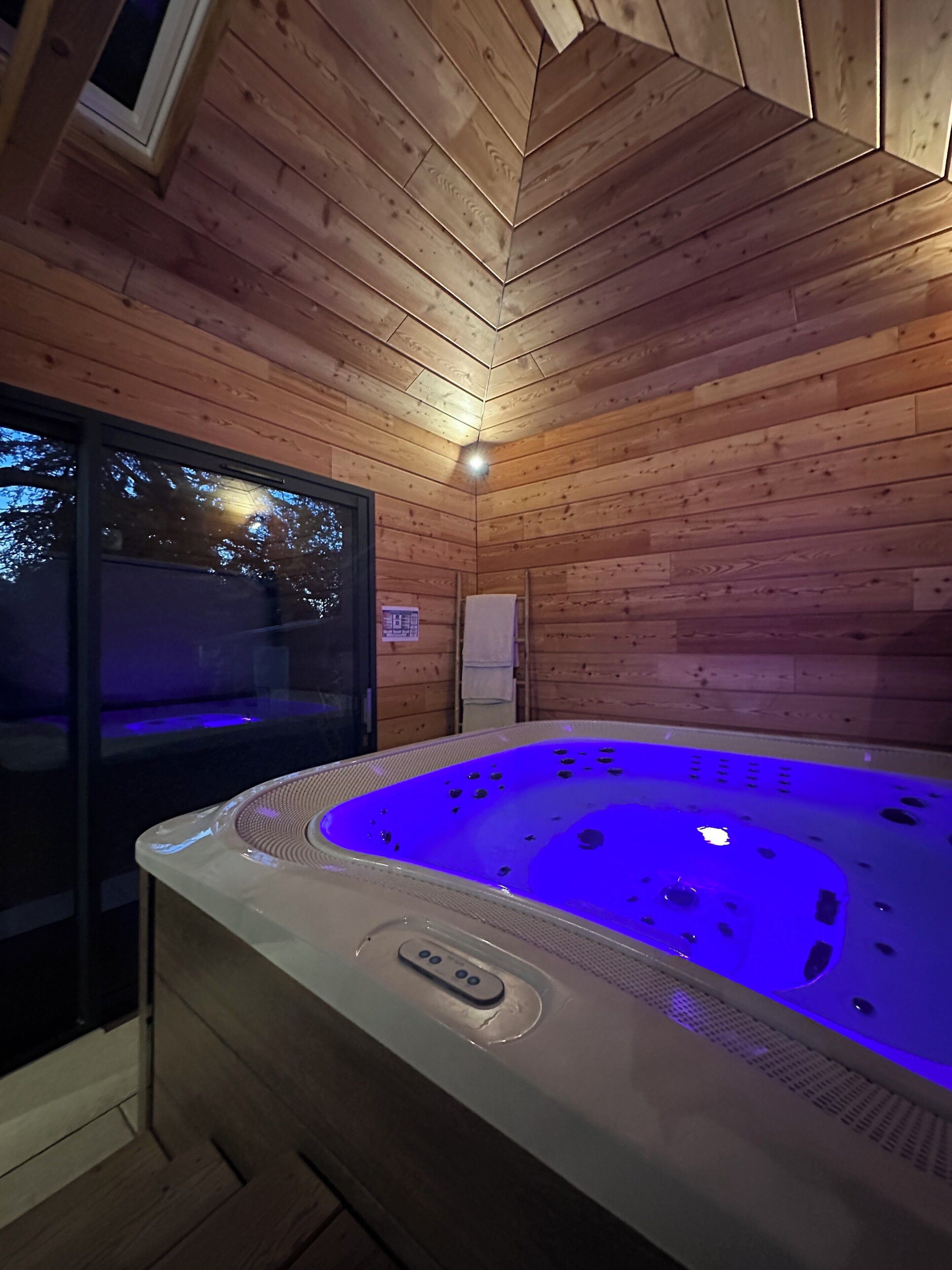 A wooden cabin with a hot tub illuminated by blue lights, a window, and a towel hanging on the wall.