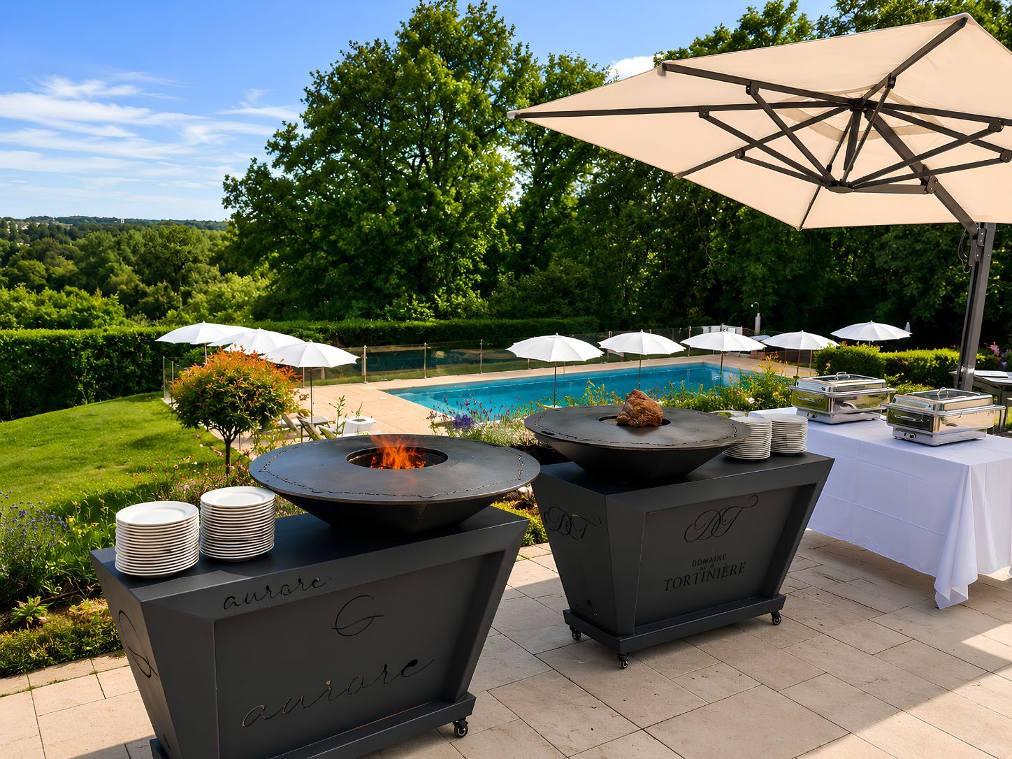 Un patio al aire libre con piscina, rodeado de follaje, que incluye una mesa con fuego, una mesa de buffet y sombrillas.