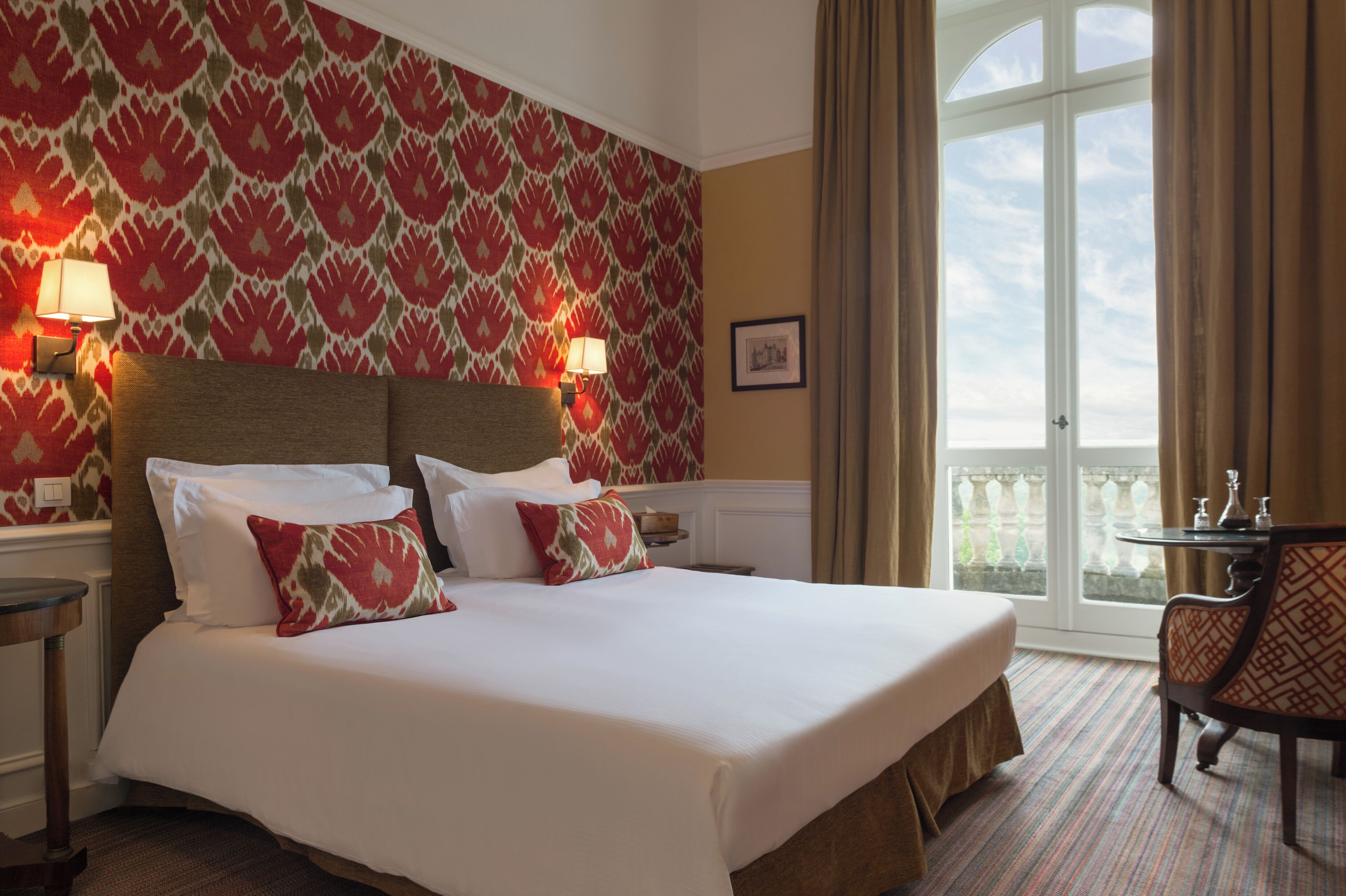A hotel room with a large bed, two bedside lamps, a framed picture on the wall, a balcony with a view, and a small table with a chair.