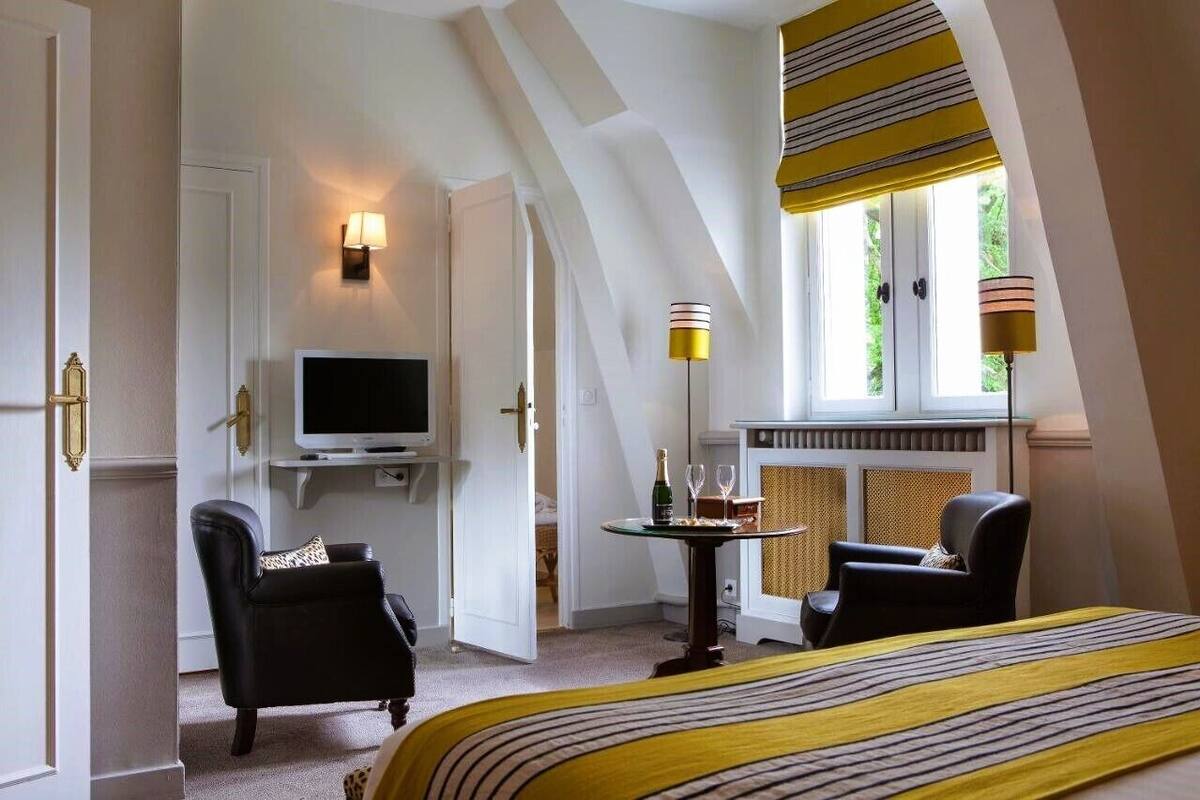 A hotel room with a bed, two chairs, a television, a table with a bottle and glasses, and a fireplace.